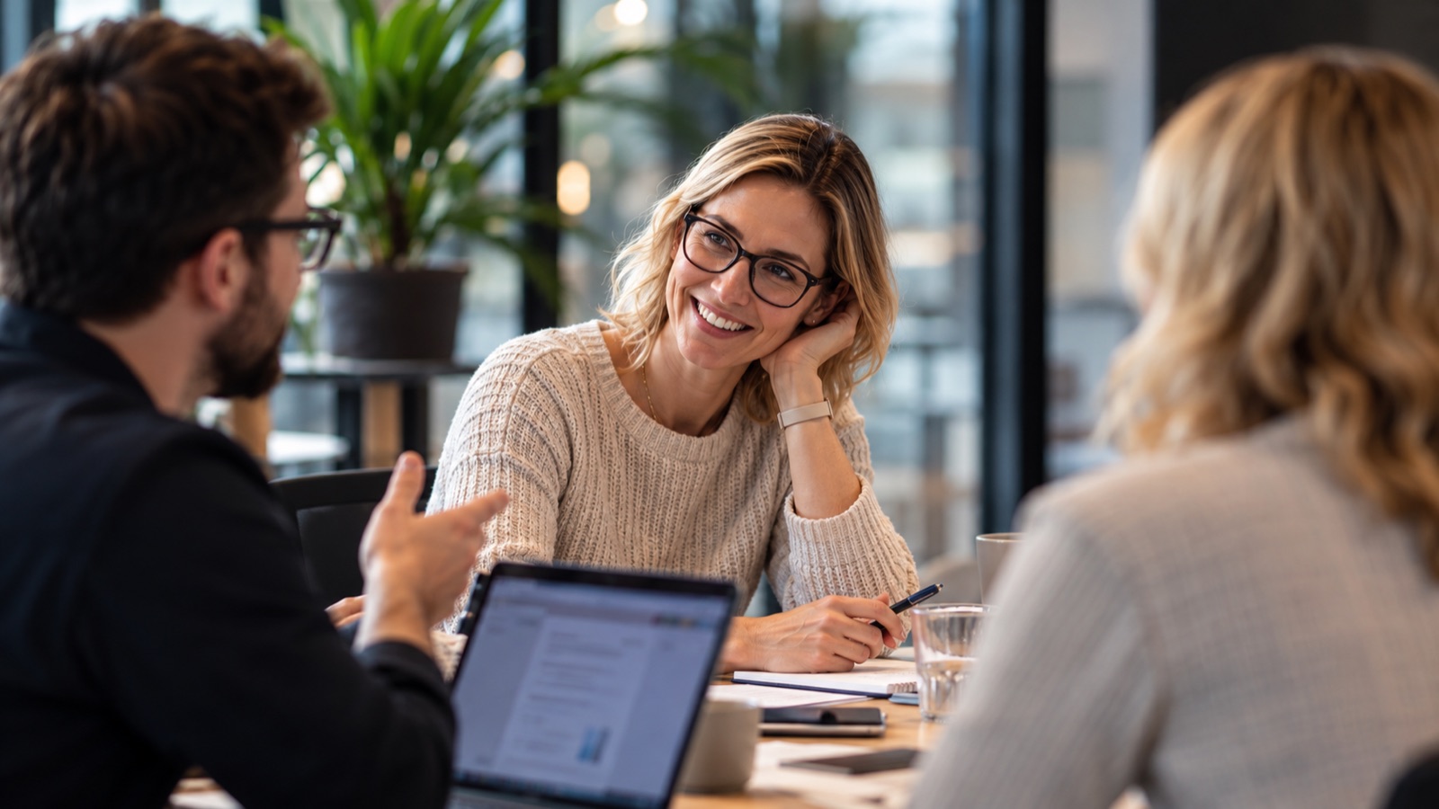 Three colleagues in conversation around a meeting table, one listening and smiling.